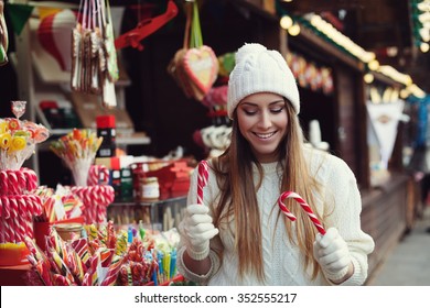 Street portrait of smiling beautiful young woman choosing candy canes on the festive Christmas fair. Lady wearing classic stylish winter knitted clothes. Close up