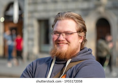 Street Portrait Of A Smiling Blond 25-30 Year Old Man With Glasses And A Beard On The Background Of The City. Perhaps He Is A Student Or An IT Specialist, A Successful Businessman Or A Salesman.