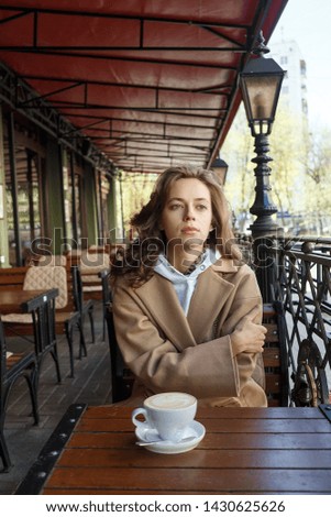 Street portrait of young woman wearing beige coat drinking coffee on a cafe veranda with dreamy and thoughtful gaze. Concept of relaxation and city leisure