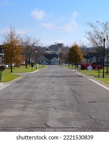 Street In A Senior Living Community. The Road Is Centered, Symmetrical, And Leads To A House At The End Of The Street. Blue Sky And The City Are Visible In The Distant Background.