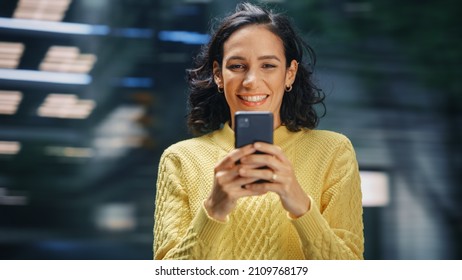 Street Shot: Portrait Of Beautiful Latin Woman Using Smartphone. Smiling Hispanic Female Entrepreneur Using Mobile Phone For Online Shopping, E-Commerce Happily. Blurred Motion Background.