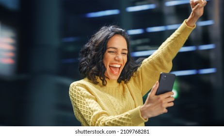 Street Shot: Portrait Of Beautiful Latin Woman Using Smartphone, Celebrating Successful Victory With Yes Gesture. Smiling Hispanic Female Entrepreneur Using Mobile Phone Happily. Blur Motion Shot.