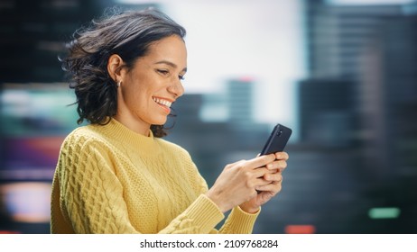 Street Shot: Portrait Of Beautiful Latin Woman Using Smartphone. Smiling Hispanic Female Entrepreneur Using Mobile Phone For Online Shopping, E-Commerce Happily. Blurred Motion Background.