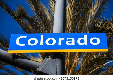 A street sign for Colorado Avenue, featuring a blue and yellow design with palm trees and a clear blue sky in the background, representing a sunny California urban environment.