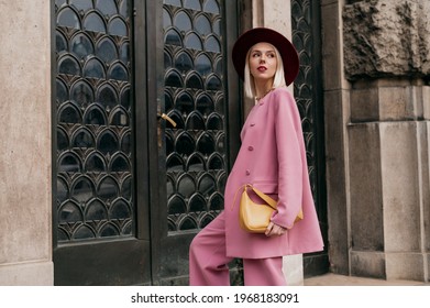 Street style, fashion conception: elegant woman wearing trendy pink suit, burgundy color hat, holding yellow bag, posing in street of city. Copy, empty space for text