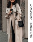 Street style fashion details of beige trench coat, woman drinking coffee against the background of the city.