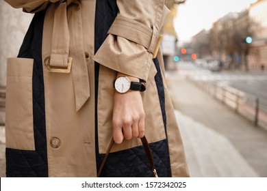 street style fashion details. close up, young fashion blogger wearing autumn trench coat and a white and golden black analog wrist watch. checking the time, holding a beautiful brown leather purse.
