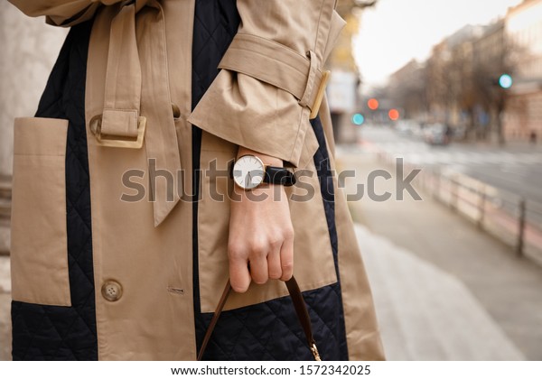 street style fashion details. close up, young\
fashion blogger wearing autumn trench coat and a white and golden\
black analog wrist watch. checking the time, holding a beautiful\
brown leather purse.\
