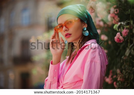 Street style fashion portrait of elegant woman  wearing trendy orange frame sunglasses, big green seashell earrings, trendy bandana, pink shirt, posing outdoor. Copy, empty space for text