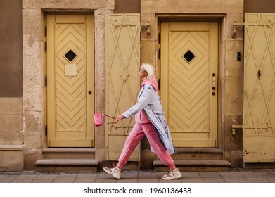 Street style, fashion: woman wearing  trendy sport chic outfit walking in street of European city. Blue trench coat, sunglasses, pink hoodie, trousers, sneakers, mini bag. Copy, empty space for text - Shutterstock ID 1960136458
