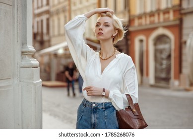 Street style photo of elegant fashionable woman wearing trendy white blouse, high waist jeans, wrist watch, with brown faux croco leather textured bag. Model walking in street of European city
