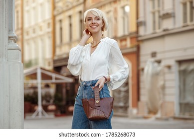 Street style photo of happy smiling fashionable woman wearing trendy white blouse, high waist jeans, holding brown faux croco leather textured bag. Model posing in street of European city. Copy space
				