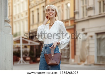 Street style photo of happy smiling fashionable woman wearing trendy white blouse, high waist jeans, holding brown faux croco leather textured bag. Model posing in street of European city. Copy space
