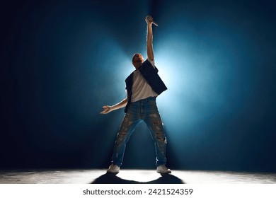 Street style singer gesturing with hand on stage against dark background. Spotlight and smoke creating silhouette effect. Concept f music and dance, lifestyle, festivals, concerts, self-expression. - Powered by Shutterstock