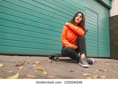 Street style teen girl. Beautiful girl skater in street clothes sitting on the street and smiling. Street portrait of a stylish girl in an orange hoodie sitting on a longboard.