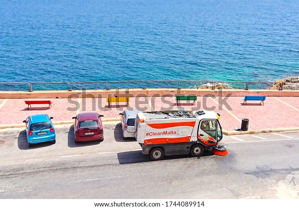 Street sweeper machine\
cleaning road near sea promenade in Malta. Wet street cleaning from\
dust in heat to improve air quality in city: Xghajra, Malta - June\
7, 2019