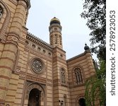 The Dohány Street Synagogue in Budapest featuring ornate Moorish architecture, yellow-red brick patterns, and golden-domed tower. Hebrew inscription above entrance shows historic Jewish heritage.