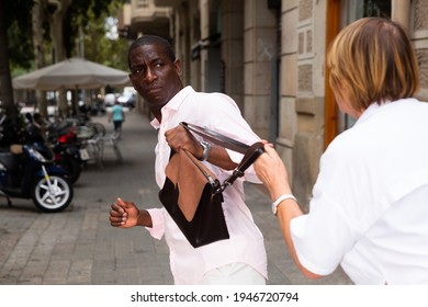 Street Thief Trying To Steal Handbag Of Scared Middle Aged Woman Walking Through City..