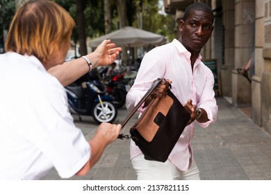 Street Thief Trying To Steal Handbag Of Scared Middle Aged Woman Walking Through City..