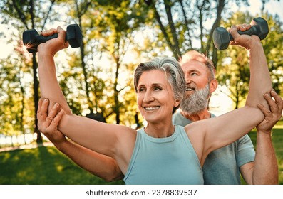 Street training together. Two joyful pensioners with happy facial expression exercising outside with sport equipment. Caring man holding woman's elbows and helping to perform exercise with dumbbells.