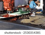 Street vendor grilling tortillas and chopped al pastor meat on a grill, in preparation of traditional tacos al pastor at a Mexican street food stand in a retail market