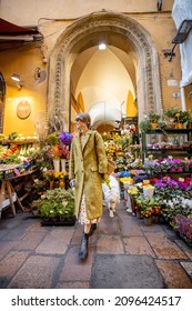 Street View On Beautiful Florist Shop With Lots Of Flowers In The Old Town Of Bologna City. Italian Woman Walking With Her Dog