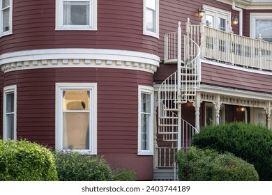 A street view of a spiral staircase on the outside of a large burgundy colored wooden house with multiple closed glass windows. Lush green shrubs are in front of the historic circular building.  - Powered by Shutterstock