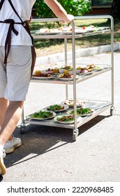 Street Waiter Carries A Three Tiered Metal Serving Trolley Cart With Various Plates Of Banquet Dishes On The Street In The Summer Under The Sun