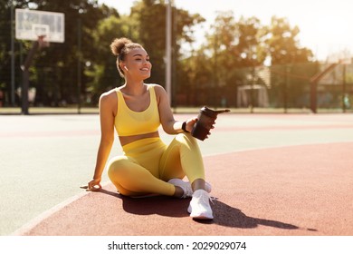 Street Workout. Pretty black lady in yellow sports clothes holding shaker bottle with water or protein resting after training on sunny day, sitting on basketball court ground, taking care of her body - Powered by Shutterstock