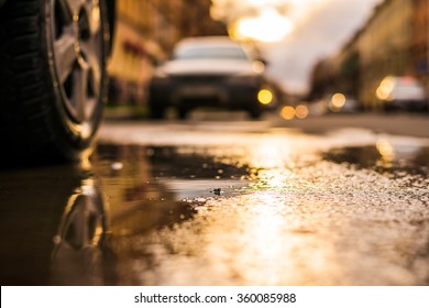 Streets In The Light Of The Autumn Sun After The Rain, View From Under The Wheels Of A Parked Car. View From Puddles On The Pavement Level