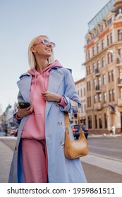 Streetstyle, Street Fashion Conception: Happy Smiling Woman Wearing Trendy Pink Sunglasses, Sport Hoodie, Trousers, Blue Trench Coat, With Yellow Fa Leather Bag, Walking In European City. 