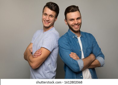 The Strength Of Brothers. Two Young Men In Casual Clothes Are Standing Back To Back With Cheerful Facial Expressions, Posing With Folded Arms And Smiling To The Camera.