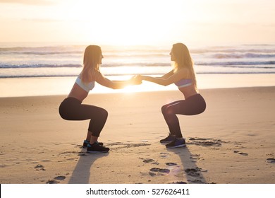 Strength in teamwork. Two young attractive female athletes exercise on the beach doing squats with a sunrise and ocean in the background. The focus is soft and dreamy.  