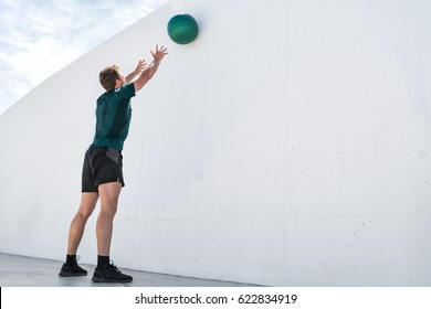 Strength Training Fit Man Cross Training Throwing Medicine Ball On Gym Wall At Fitness Centre. Workout Bodyweight Exercises Using Heavy Weight Ball. Overhead Medicine Ball Throw Plyometric Exercise.