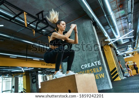 Strength training. Full length young athletic woman in sportswear jumping on wooden crossfit box at gym