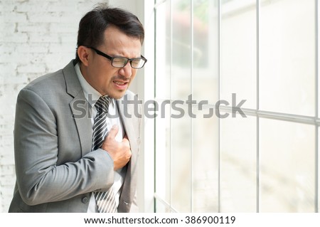 Stress Asian Businessman standing near the window and having chest pain. Man wear eyeglasses having heart attack or failure during working at office. Exhausted, tired, ill from overworked. 