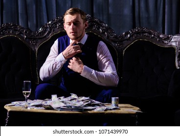 Stress and business concept. Cash, money, banknotes on table. Man in waistcoat, businessman sit in dark luxury interior background. Macho with stressed face untie tie, relax after business meeting.