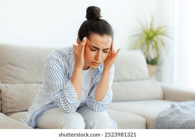 Stress, headache, illness. Upset mixed race woman sitting on a sofa in living room with eyes closed, suffering headache, vertigo, migraine, having cramp in a templess, need treatment and rest - Powered by Shutterstock