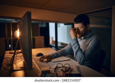 Stress, night and business man at desk by computer for fatigue, overtime or project deadline in office. Tired, publishing agency and employee for exhausted, reading online report or decision in dark - Powered by Shutterstock