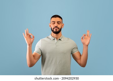 Stress relief concept. Middle eastern man meditating with closed eyes, keeping calm on blue studio background. Young arab guy practicing yoga, searching for inner balance