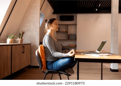 Stress relief during freelance work. Brunette woman freelancer sitting in lotus pose and meditating with hands in mudra gesture on chair in front of open laptop on table, working from home - Powered by Shutterstock