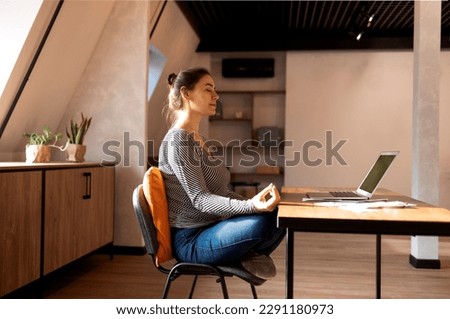 Stress relief during freelance work. Brunette woman freelancer sitting in lotus pose and meditating with hands in mudra gesture on chair in front of open laptop on table, working from home