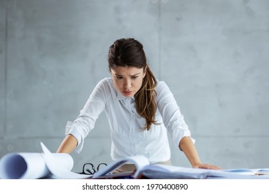 Stress At Work. Busy Female Architect Leaning Against A Desk And Looking At Blueprints And Papers. Young And Ambitious Woman Has A Worried Expression And She Is Not Happy With How The Project Is Going