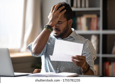 Stressed african businessman sit at desk hold paper letter reading bad awful negative news got fired from work. Dismissal notice, financial problems debt notification from bank, unpaid invoice concept