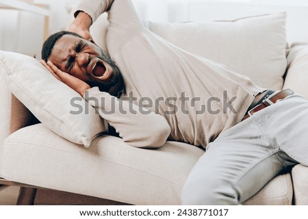 Stressed AfricanAmerican man with a headache sitting alone on the couch at home His tired and sick expression reflects the physical and mental pain he is experiencing The living room's background adds