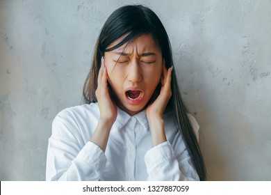 Stressed Angry Stubborn Beautiful Asian Girl In White Shirt Sticking Plug Fingers In Ears Screaming Loud Feel Annoyed, Mad Crazy Rebellious Teen Girl Yelling In Tantrum  On Grey Studio Background.