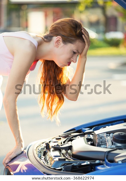 Stressed, angry\
young woman in front of her broken down car with opened hood\
looking at engine, outside street, road. Negative face expressions,\
emotions, feelings. Bad luck, lemon\
car