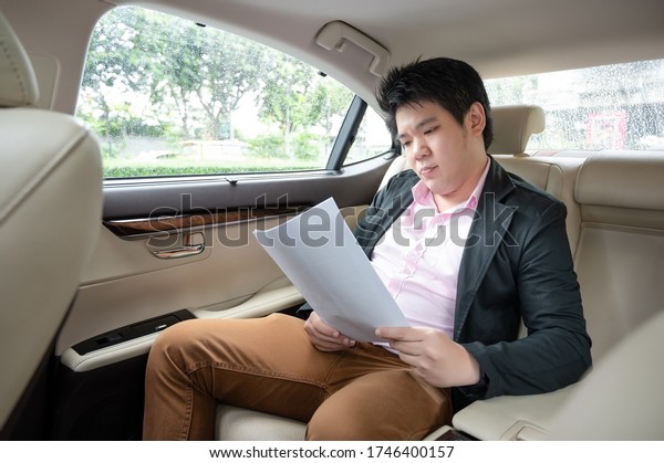 Stressed asian businessman looking\
at Financial documents with expression Tired and worried while\
sitting on the back seat in the car. many bad financial\
report.