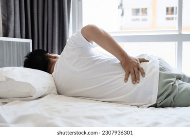 Stressed asian man having a backache,sore hips,waist hurts,unhappy adult people suffering from low lumbar pain or acute back strain,lying on the mattress at home,health care,medical,lifestyle concept