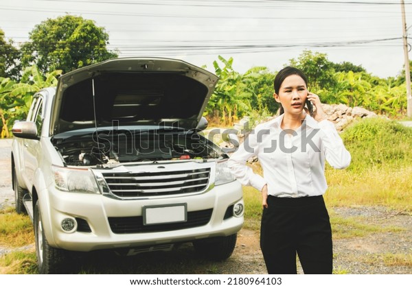 Stressed asian woman stands in front broken car\
on her commute to work, using her smartphone to call an online call\
center asking for urgent emergency help urgent from an insurance\
company.